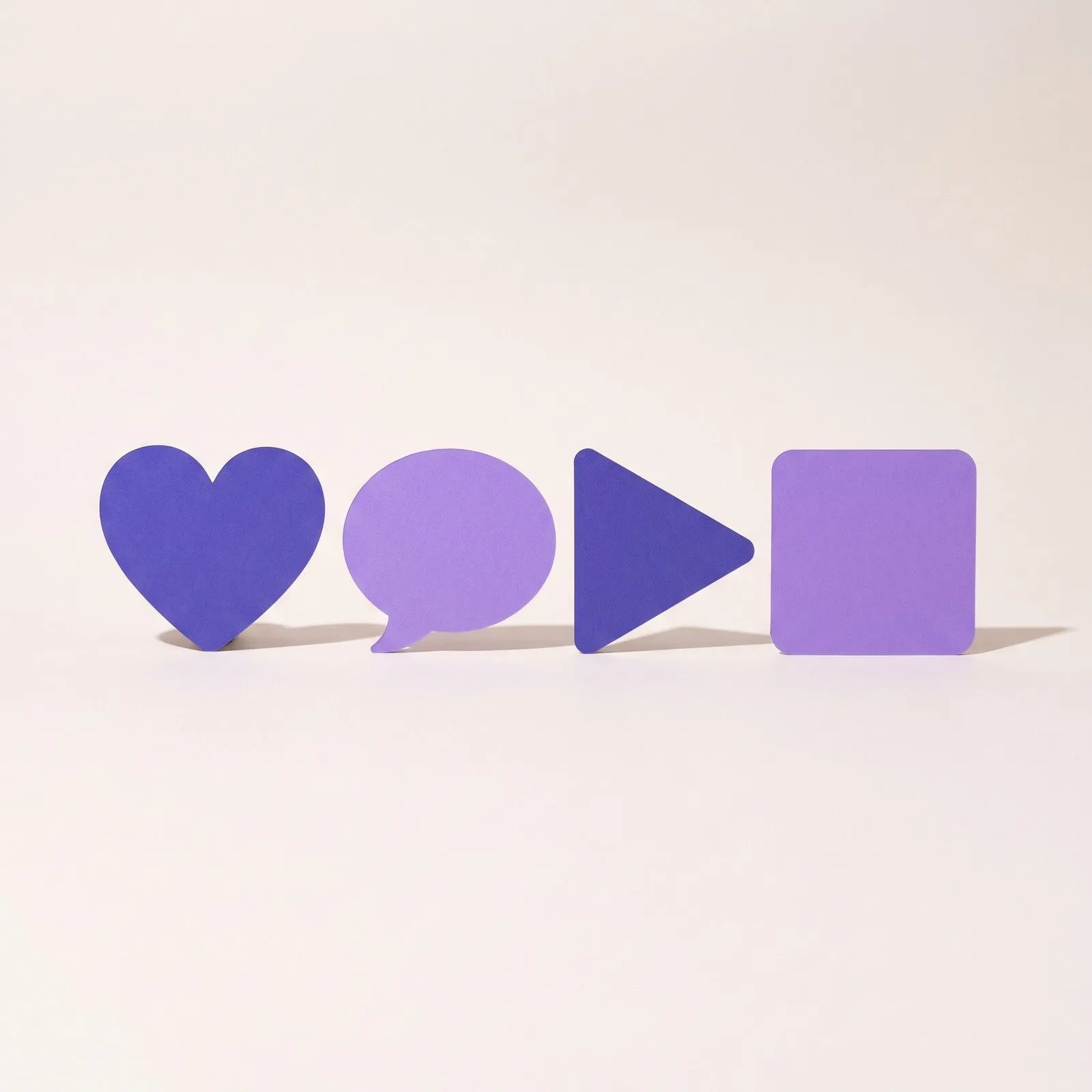 Vier verschieden große Papierformen in Violett nebeneinander: Herz, Sprechblase, Play-Dreieck und Quadrat