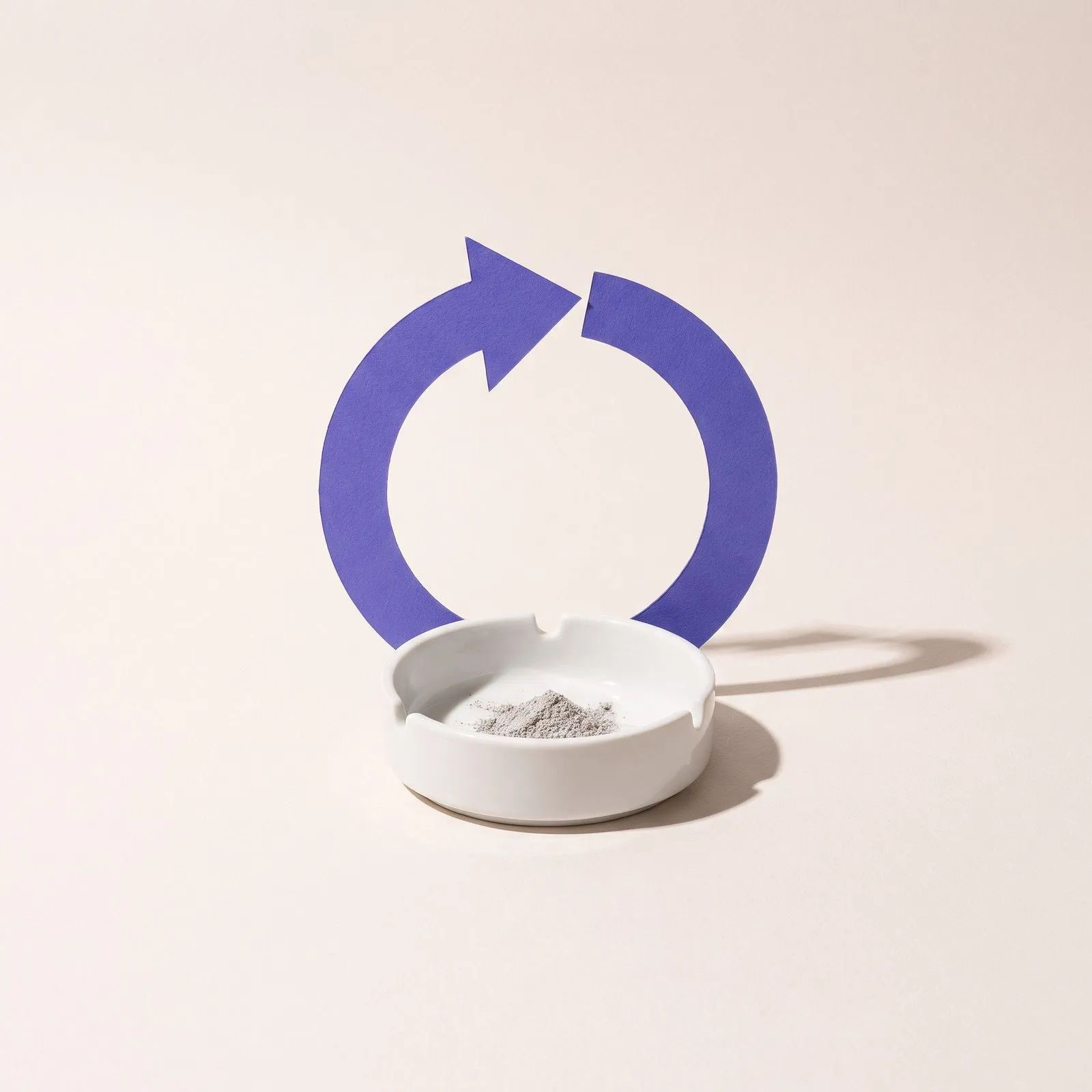 Weißer Aschenbecher mit etwas Asche, dahinter ein violetter Kreispfeil aus Papier